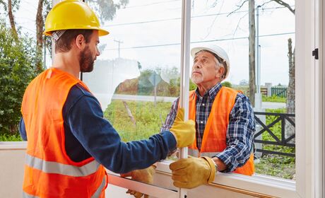 Bild Zwei Bauarbeiter mit Helmen und Warnwesten installieren gemeinsam ein Fenster in einem Gebäude.