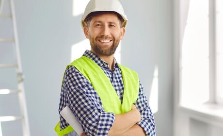 Bild Ein lächelnder Projektleiter steht in einem Raum mit Fenstern, trägt einen Bauhelm und hält Baupläne in der Hand.